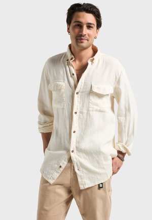 Hombre con camisa color crema abotonada y pantalones beige, de pie con las manos en los bolsillos contra un fondo claro y uniforme.