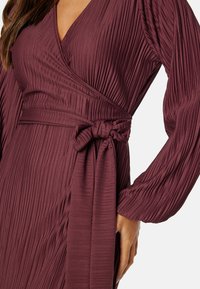 Bordowa, plisowana sukienka z długimi rękawami i krojem kopertowym, z wiązanym paskiem w talii. Gładka tekstura i głęboki dekolt w kształcie litery V.