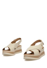 Sandalias de cuña de cuero crema con diseño de tiras cruzadas, hebilla ajustable y una suela exterior de color marrón claro en contraste. Textura suave y punta abierta.