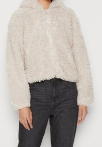 Personne portant une veste zippée beige clair et duveteuse à manches longues, avec un jean noir taille haute, sur un fond uni.