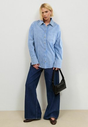 Femme aux cheveux blonds courts portant une chemise bleu clair oversize, un jean sombre à jambes larges, des chaussures marron et tenant un petit sac à main noir.