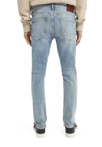 Jeans de denim azul claro con un corte slim, que cuentan con bolsillos traseros, detalles de desgaste y una etiqueta de piel marrón en la cintura. Llevados con zapatillas negras.