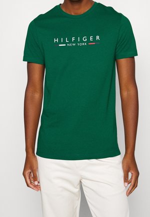 Mężczyzna ubrany w zieloną koszulkę z krótkim rękawem z napisem "HILFIGER NEW YORK" w kolorze białym, połączoną z białymi spodniami.