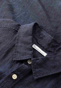 Marineblaues Hemd mit Knöpfen aus strukturiertem Material, das einen Kragen, weiße Knöpfe und ein Pflegeetikett mit der Aufschrift "Knowledge Cotton Apparel" aufweist.