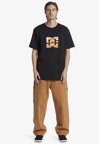 Camiseta de algodón negra con un logotipo gráfico dorado en la parte delantera, combinada con pantalones cargo sueltos color beige y zapatillas negras.