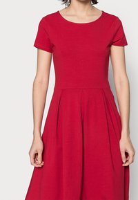 Czerwona sukienka z krótkim rękawem, z dekoltem w serek, dopasowaną górą i rozkloszowaną spódnicą z plisami. Wykonana z miękkiej bawełnianej tkaniny.