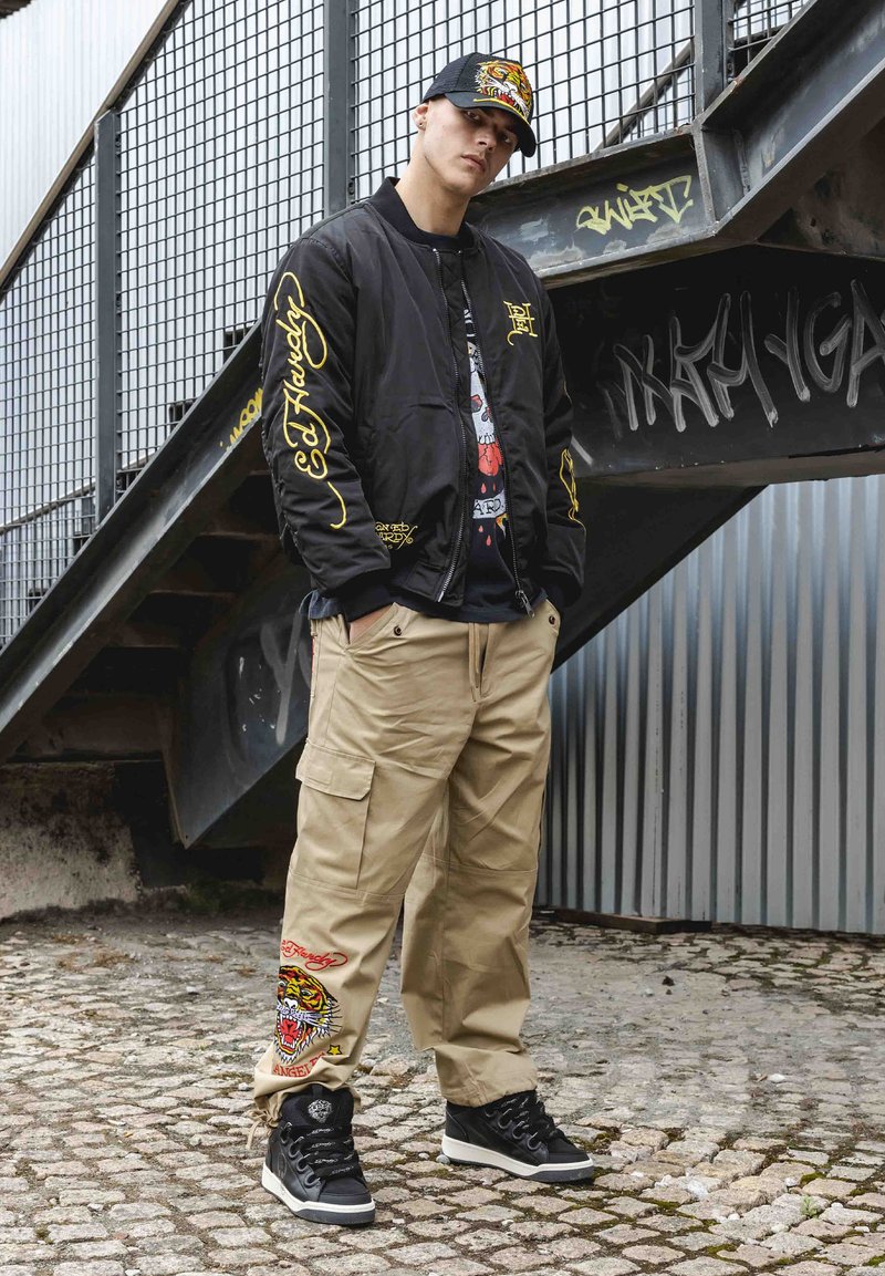 Svart bomberjacka med gul lettering, grafisk t-shirt, khaki cargobyxor med tigermönster och svarta hög-top sneakers. Urban miljö.