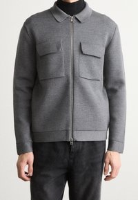 Šedá bunda na zip z texturované látky, s límcem, dvěma náprsními kapsami a žebrovanými manžetami a lemem. Nošena přes černý top.