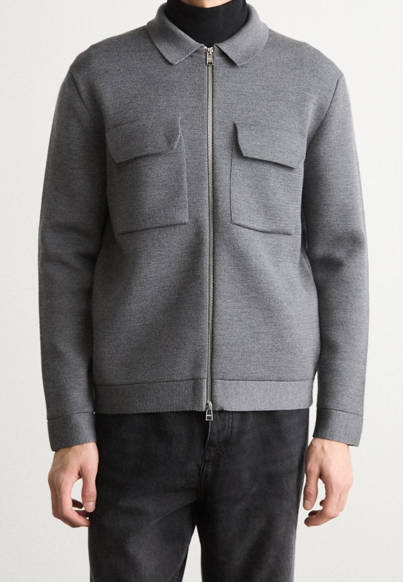 Šedá bunda na zip z texturované látky, s límcem, dvěma náprsními kapsami a žebrovanými manžetami a lemem. Nošena přes černý top.