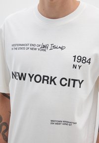 Camiseta de algodón blanca con grandes gráficos de texto negro que dicen "NUEVA YORK", "1984" y varios monumentos y frases de Nueva York.