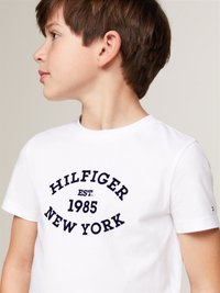 T-shirt en coton blanc avec un texte imprimé bleu marine : "HILFIGER EST. 1985 NEW YORK." Manches courtes, col rond, design simple.