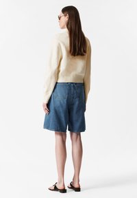 Cremfarbener Strickpullover mit geripptem Saum, kombiniert mit blauen Jeansshorts in lockerer Passform und zwei Rückentaschen. Schlichte Sandalen runden den Look ab.