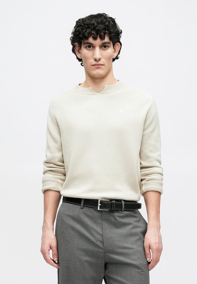 Ανοιχτό μπεζ πλεκτό πουλόβερ με μοναδικό γιακά, συνδυασμένο με γκρι παντελόνια και μαύρη ζώνη, με διακριτικές ραφές.