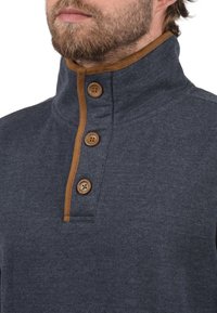 Marineblauwe pullover met een hoge kraag, voorzien van bruine knoopaccenten en een gedeeltelijke sluiting. Zachte textuur met een casual ontwerp.