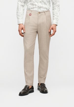 Hombre con pantalones de vestir beige, camisa blanca con estampado metida por dentro y mocasines de cuero negro, de pie sobre fondo blanco.