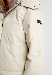 Veste d'hiver beige matelassée avec fermeture éclair et boutons-pression, dotée d'un col intérieur gris et d'un écusson noir "ROXY" sur la manche.