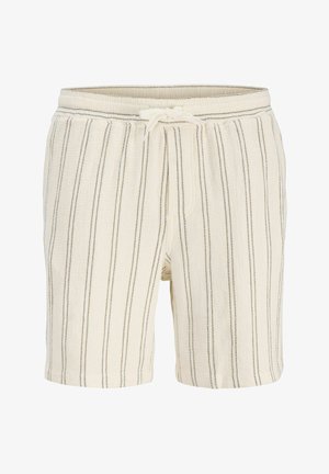 Nastolatek z kręconymi włosami, ubrany w kremową koszulkę w paski z krótkim rękawem i dopasowane szorty, białe skarpetki, trampki oraz czerwoną apaszkę na szyi.