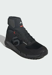 Czarny but sportowy z siateczkową cholewką, wyściełanym wsparciem kostki i teksturowaną podeszwą. Akcenty obejmują czerwoną naszywkę oraz logo po boku.