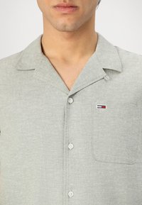 Lichtgrijs kortmouwen shirt van textuurstof. Kenmerkt zich door een ingesneden kraag, een voorknoopzakje en een klein geborduurd logo op het zakje.