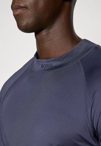 Mørkeblå træningsskjorte med en glat tekstur, der har en høj krave og en sort "BORG" logo ved halsudskæringen. Iøjnefaldende sømdetaljer.