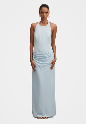 Žena v svetlomodrých šatách bez rukávov so zaviazaním za krk a riaseným detailom v páse, zlaté kruhové náušnice a ploché sandále.