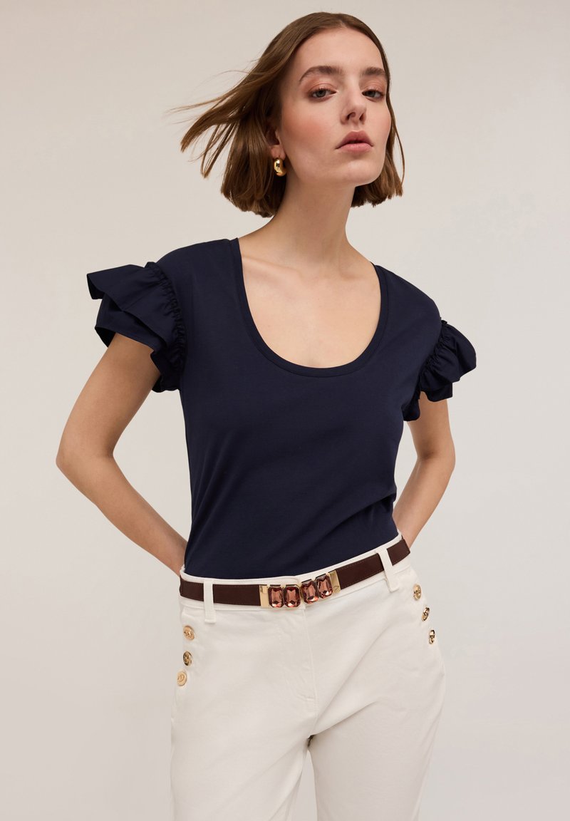 T-shirt blu navy con maniche corte a volant; scollatura rotonda. Abbinato a pantaloni bianchi con dettagli di bottoni dorati e una cintura marrone.