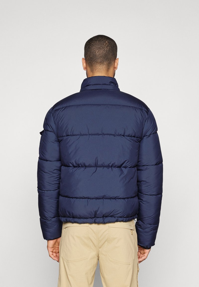 Marineblaue Steppjacke mit horizontaler Steppung, hohem Kragen und elastischen Bündchen. Entworfen mit einem matten Finish und ohne sichtbare Beschläge.