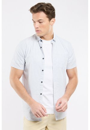 Jeune homme portant une chemise légère à manches courtes bleu clair par-dessus un t-shirt blanc et un pantalon beige, regardant sur le côté devant un fond uni.