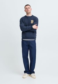 Marineblaues Sweatshirt mit einem strukturierten Buchstaben "B"-Patch, kombiniert mit blauen Jeanshosen und cremefarbenen Schnürschuhen. Schlichte, lockere Passform mit Rundhalsausschnitt.