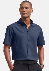 Jūras spēku īsām piedurknēm krekls ar pogām, izgatavots no teksturētas auduma, ar izplatītu apkakli un divām priekšējām pogām, kombinēts ar melnām biksēm un jostu.