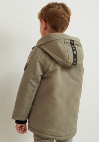 Coat in light beige, featuring a hood, black accents, and a rounded hem. Side pockets and a textured fabric complete the design.