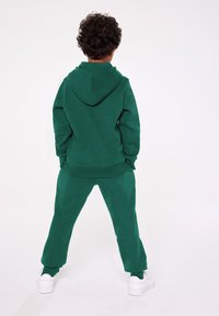 Grön huvtröja och matchande sweatpants i mjukt material. Huvtröjan har snörning och ficka, med en avslappnad passform. Vita skor.