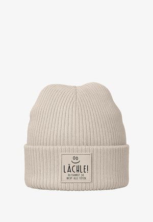 Light beige knitted beanie with a ribbed texture and folded cuff. Features a rectangular label with black text and a smiley face detail.