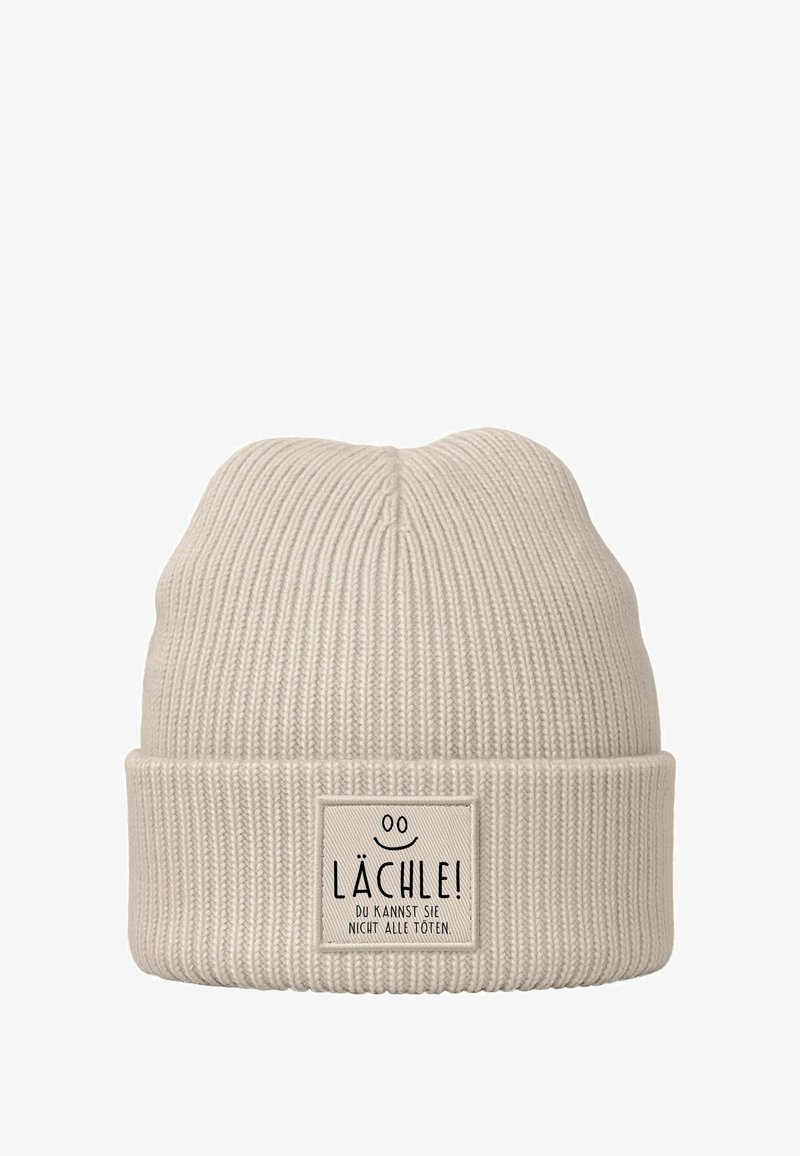 Light beige knitted beanie with a ribbed texture and folded cuff. Features a rectangular label with black text and a smiley face detail.