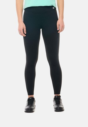 Personne portant des leggings noirs ajustés, des chaussures de sport noires et un haut vert clair, debout devant un fond blanc uni.