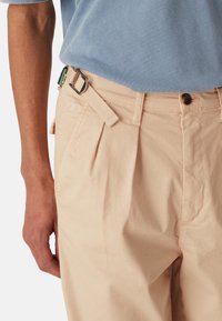 Muž ve světlých plisovaných kalhotách s látkovým pásem a modré košili, ruka odpočívající vedle kapsy na bílém pozadí.