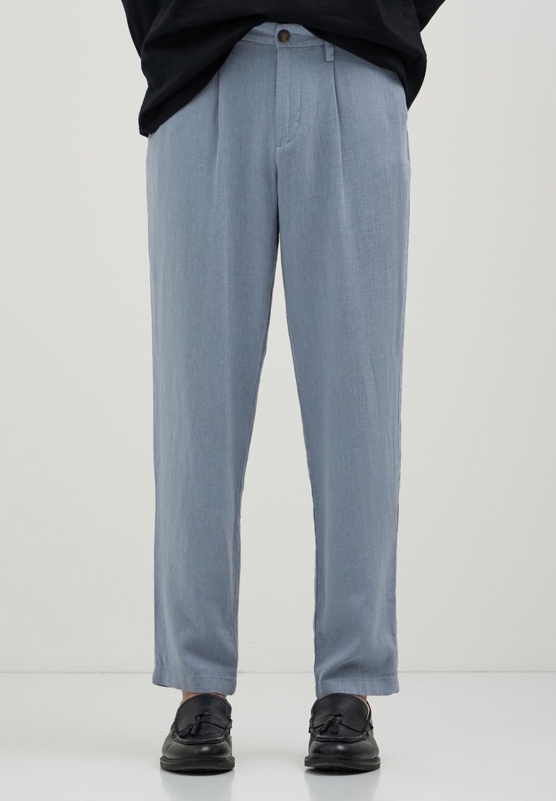 Pantalon plissé bleu clair associé à des mocassins en cuir noir à pampilles sur une personne debout devant un fond uni.