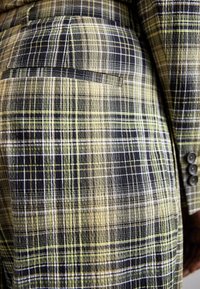 Gros plan d'un tissu à carreaux noir, beige et vert sur une veste et un pantalon, montrant une poche arrière et une manche de veste avec des boutons.