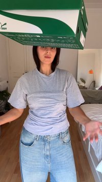 Caixa de sapatilhas verde da Puma segurada acima de uma pessoa que veste uma t-shirt cinza e jeans azul claro. A t-shirt está ajustada à cintura, criando textura.