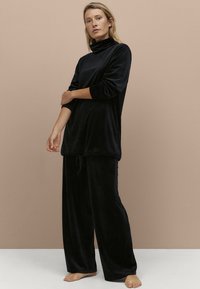 Černý sametový loungewear set s vysokým límcem, dlouhými rukávy a kalhotami s nastavitelným šňůrkovým pasem, hladká textura a uvolněný střih.