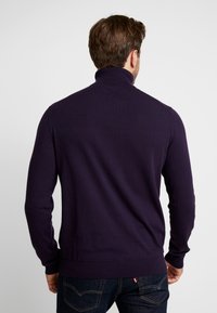 Dunkellila Pullover mit hohem Kragen, gerippten Bündchen und Saum; hat eine glatte Textur und eine taillierte Passform und wird von einer Person getragen, die sich abgewandt hat.