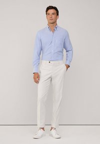 Camisa de botón azul claro con un solo bolsillo en el pecho y pantalones blancos, que presenta un corte ajustado y detalles en los botones marrones, combinada con zapatillas blancas.