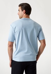 Tricou scurt cu mânecă în culoare albastru deschis din material textil de bumbac, cu guler rotund și croială lejeră, având o textură netedă și cusături minime.
