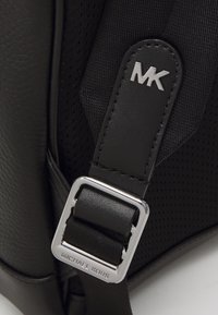 Μαύρη δερμάτινη και δίχτυνη τιράντα σακιδίου με ασημένιο μεταλλικό κούμπωμα. Διαθέτει ανάγλυφο λογότυπο και η ομαλή υφή αντιπαραβάλλεται με το τρυπημένο υλικό.