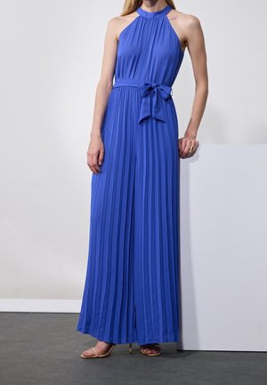 Žena v bezrukávých, královsky modrých plisovaných maxi šatech s halterovým výstřihem a zavázaným pasem stojí s jednou rukou opřenou o bílý povrch.
