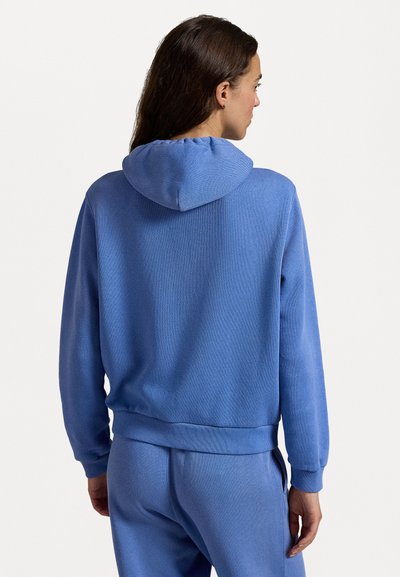 Blauer Kapuzenpullover, gestrickte Textur, gerippte Bündchen und Saum, lockere Passform, mit einem gerafften Kapuzendetail, getragen mit passenden Hosen.