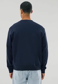 O persoană stă cu spatele, purtând un hanorac bleumarin și blugi deschiși la culoare, pe un fundal simplu deschis.