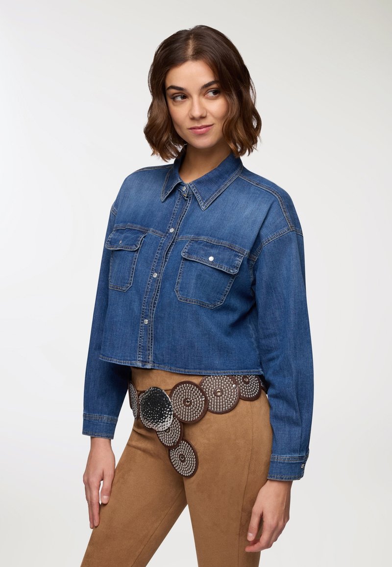 Giacca di jeans tagliata in blu medio con due tasche sul petto, chiusura a bottoni e maniche lunghe. Indossata con pantaloni marroni e una cintura decorativa.