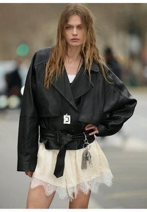 Mujer con cabello largo y ondulado que lleva una chaqueta de cuero negra extragrande, falda blanca con encaje en los bordes y accesorios plateados de llavero, posando al aire libre.