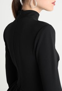 Musta korkeakauluksinen mekko, jossa on pitkät hihat, muotoiltu vartalo-osa, pehmeä kangas ja piilotettu vetoketju takana.